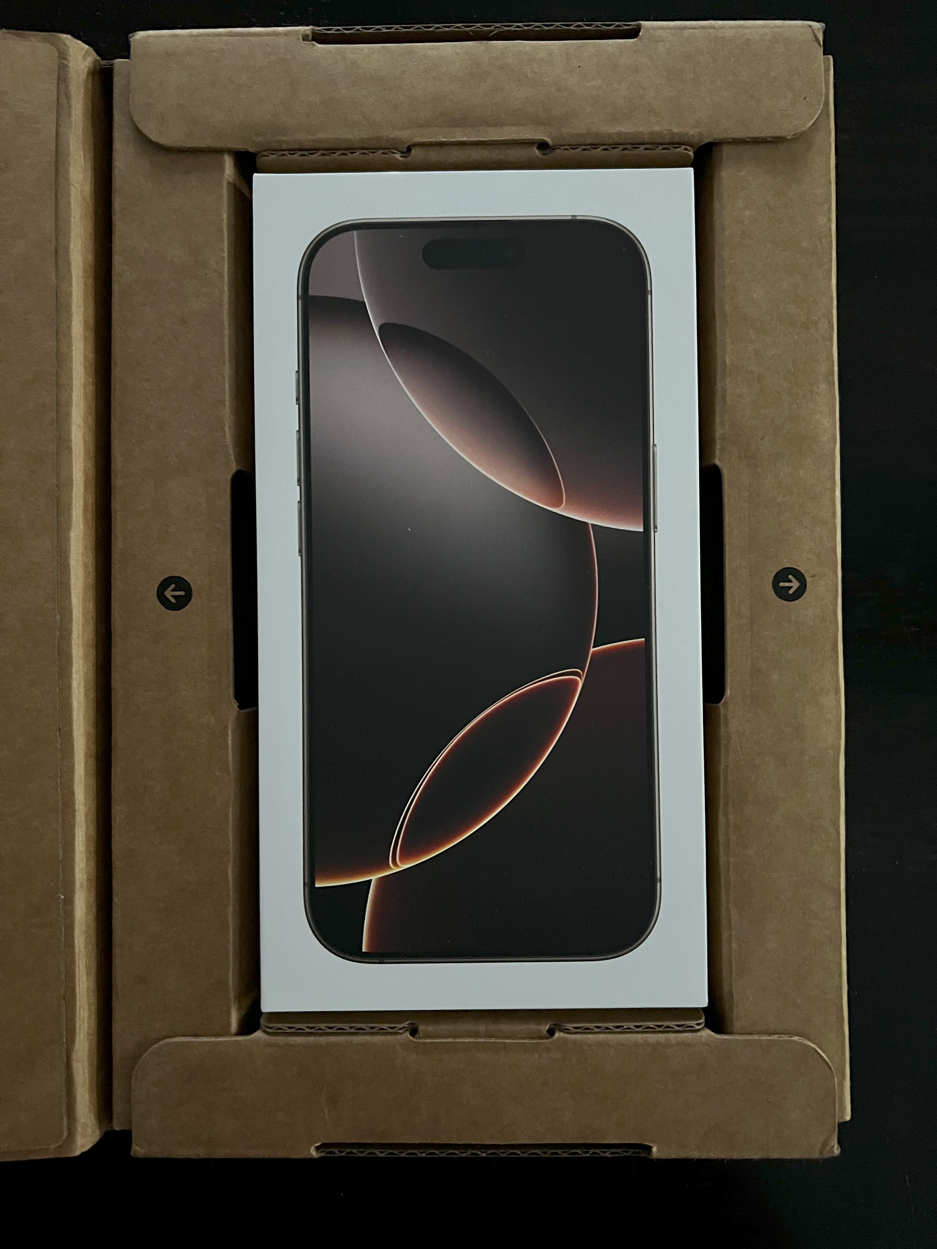 An unopened iPhone 16 Pro box lays in its cardboard coffin/transport vessel.